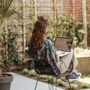 Vrouw werkt buiten met laptop op schoot, zittend op een terras in de tuin.