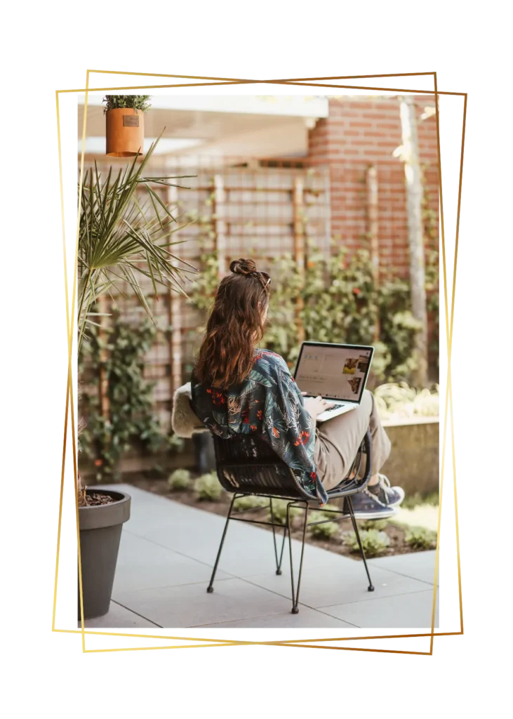 Vrouw werkt buiten met laptop op schoot, zittend op een terras in de tuin.