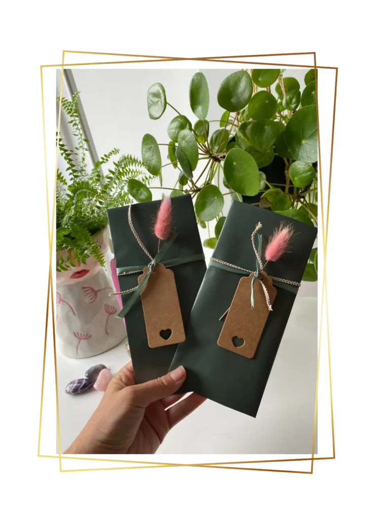 Hand houdt twee ingepakte cadeaubonnen met label en roze pluimpje, tussen groene planten.
